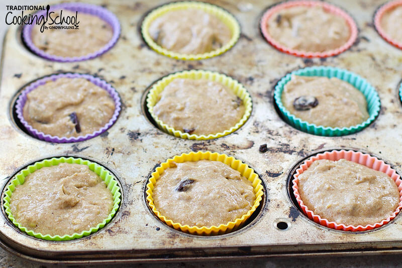 a muffin tin of brightly colored silicone muffin cups filled mostly to the top with muffin batter in which raisins are visible