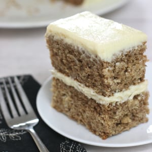 A double layered sourdough spice cake with cream cheese frosting between layers and on top sitting on a small white plate with a form beside it.