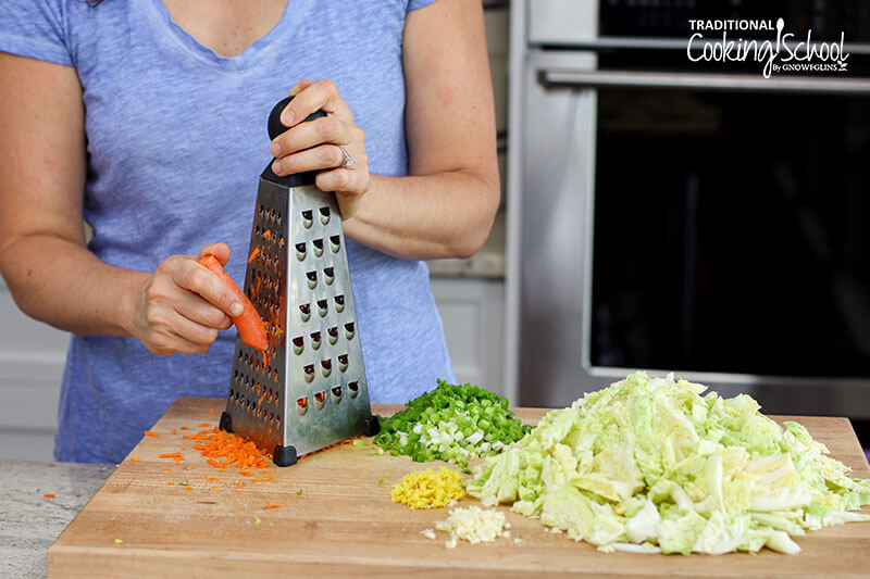 Woman in a blue shirt grating carrots onto a cutting board that has chopped garlic, ginger, cabbage and green onions for a homemade Kimchi recipe.
