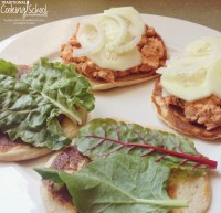 Two salmon salad sandwiches on sourdough english muffins topped with salad greens and mayo on a large white plate.