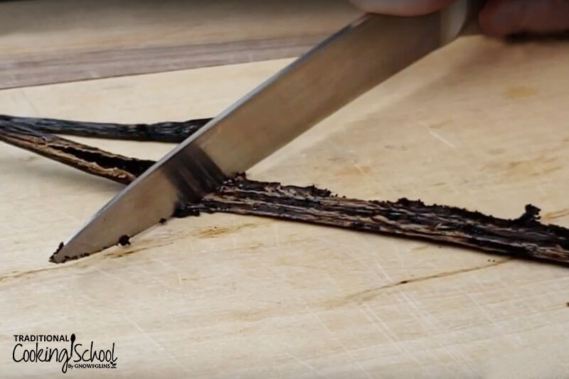 A vanilla bean on a cutting board with a paring knife scraping out the vanilla bean seeds.