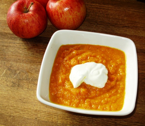 bright orange soup garnished with a dollop of sour cream next to two whole apples