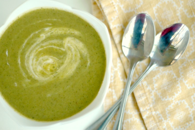 fresh green colored soup swirled with white next to a pale orange napkin and two silver spoons