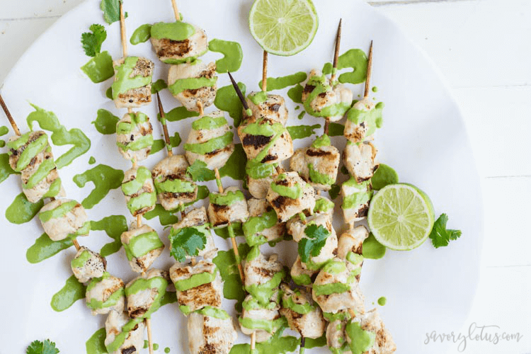 Cilantro sauce topped chicken kabobs with lime halves on white plate