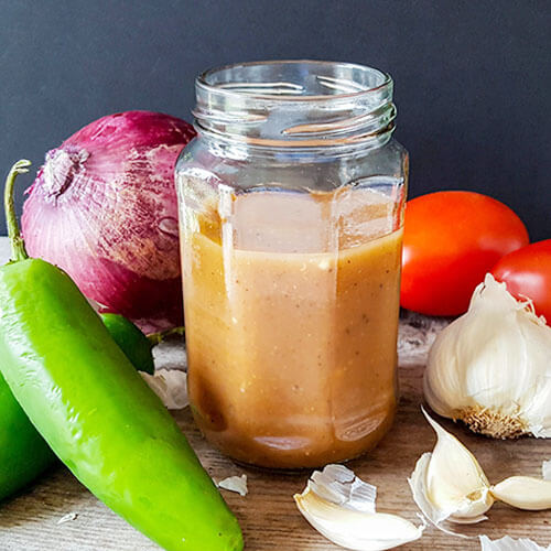 Homemade hot sauce in a small jar surrounded by garlic, hot peppers, onion, and tomatoes.