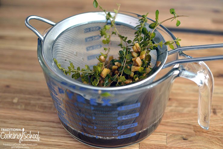 Image of a glass measuring cup with a strainer basket sitting on top with herbs and spices strained out and tea for homemade cough syrup in the measuring cup.