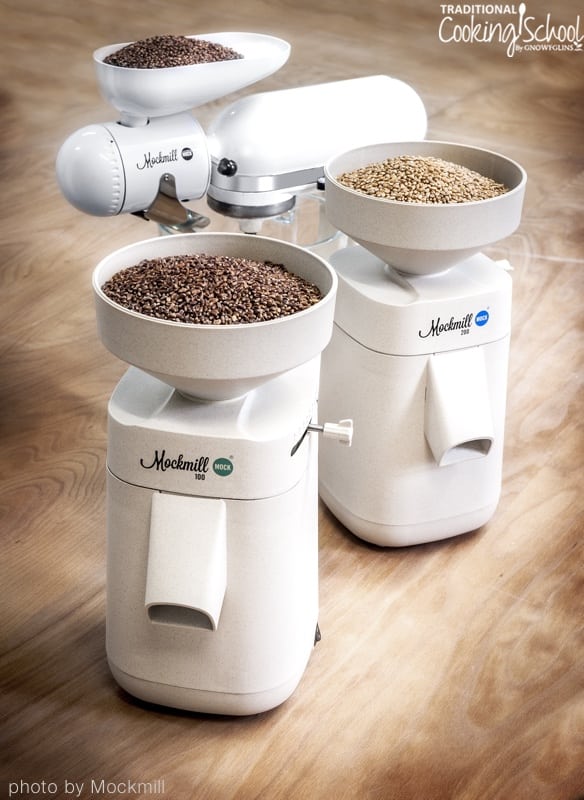 Two models of Mockmill and the Mockmill Kitchenaid attachment on a wooden surface with grain berries ready to be ground into flour.