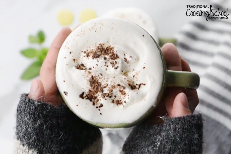 white hot chocolate in cup with whipped cream, cinnamon, and held by hand with long sweater