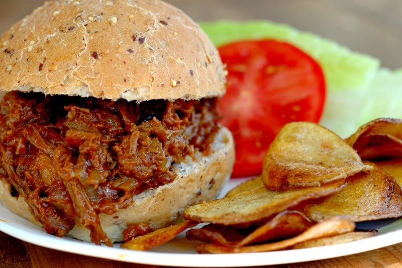 Shredded beef BBQ sandwich with a side of homemade chips, lettuce, and tomato on white plate