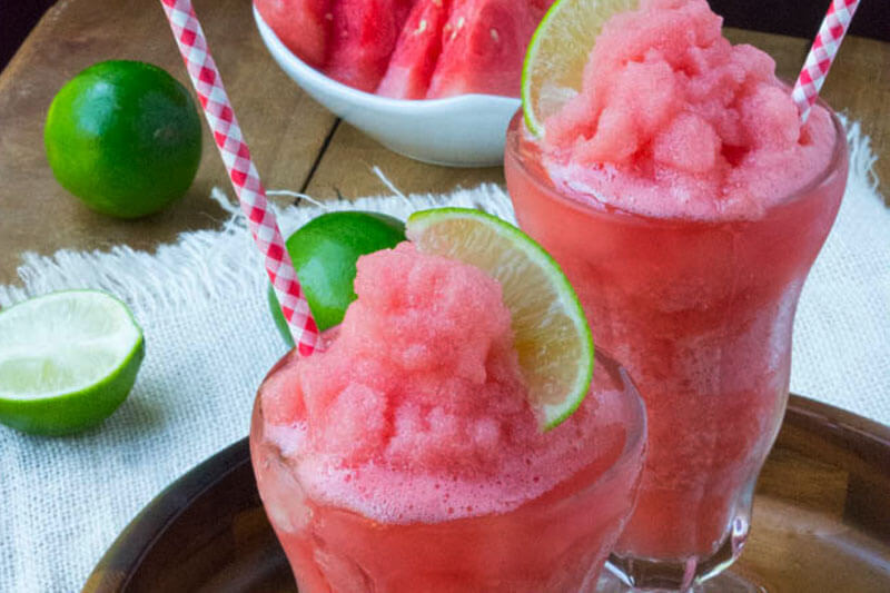 frozen watermelon smoothies in fountain glass on wooden platter surrounded by fresh watermelon and limes
