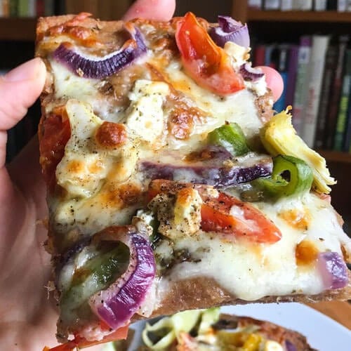 hand holding up a piece of mediterranean sourdough pizza topped with cheeses, tomatoes, bell peppers, onions, and artichoke hearts