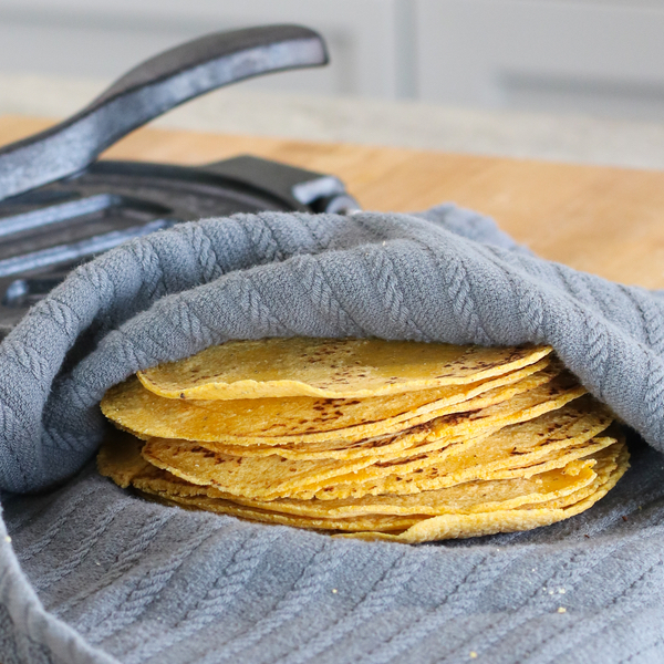 stack of authentic corn tortillas wrapped in a gray cloth with a tortilla press in the background