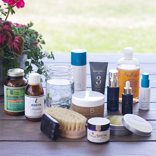 array of many different non-toxic products on a picnic table, including cod liver oil, tallow balm, Shea Moisture shampoo, BeautyCounter products, a dry brush, etc.