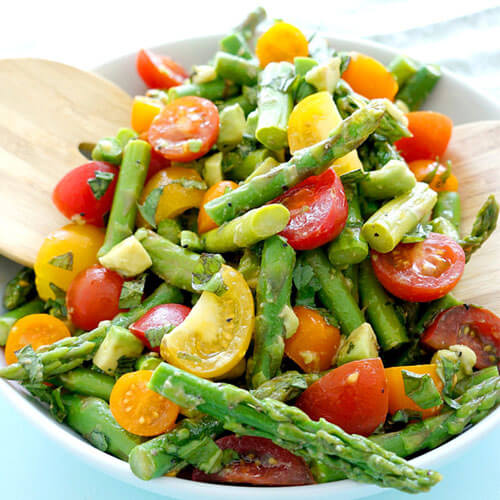blue bowl of asparagus tomato salad with wooden serving tongs