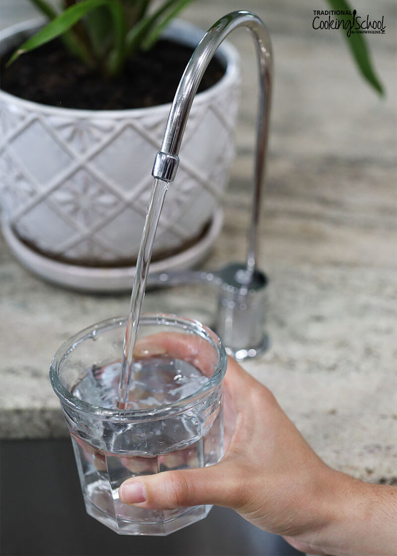 woman's hand holding a transparent glass of water as it fills from a water filter faucet at the sink