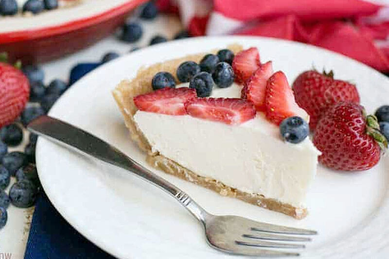 slice of low carb, no bake cheesecake on a plate next to a fork and several whole strawberries, with blueberries and sliced strawberries on top
