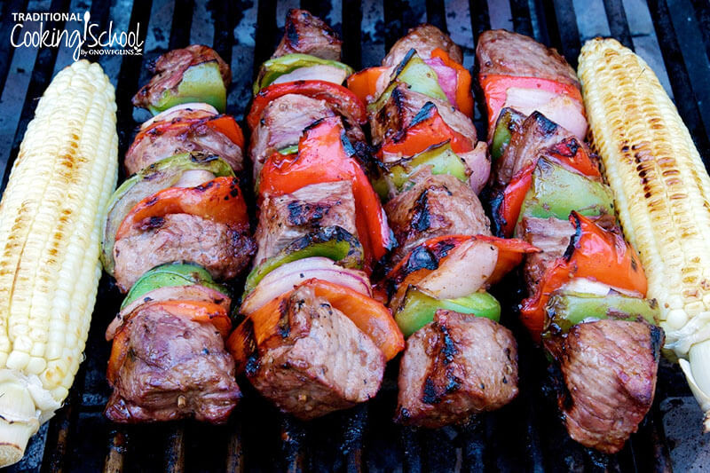 four shish kabobs of bell peppers, onion, and meat chunks on a grill flanked by two ears of corn