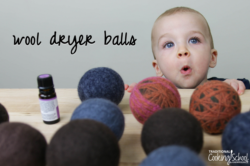 homemade wool dryer balls in a variety of colors (mostly brown and blue) on a wooden table top next to a bottle of lavender essential oil, with a small blue-eyed boy peering over the edge of the table looking surprised, with text overlay: "wool dryer balls"