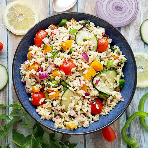 overhead shot of a dark blue ceramic bowl of cold brown rice salad with garden veggies like cherry tomatoes, sliced cucumber, bell pepper, and diced onion, on a wooden backdrop surrounded by prettily sliced veggies