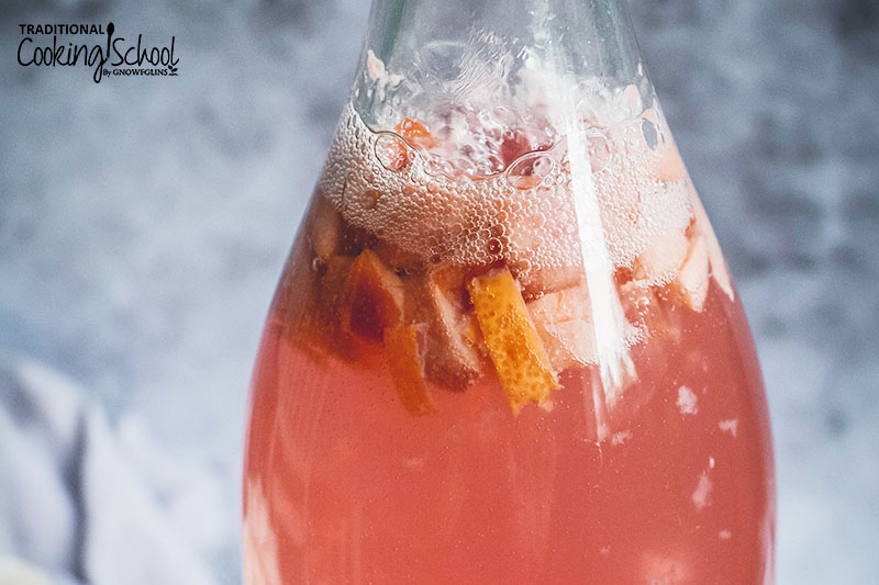 close-up shot of the narrow neck of a flip-top glass bottle full of light pink water kefir that is fizzing around small pieces of lemon and cherry