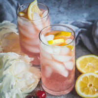 two clear drinking glasses full of a ice cubes and a fizzy light pink liquid, topped with lemon slices, and surrounded by white flowers, cherries, and lemon halves