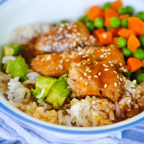 blue and white ceramic bowl of chicken pieces covered in a honey colored glaze with sesame seeds sprinkled over top, next to avocado chunks, small carrot cubes, and peas, on a bed of rice