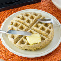 white plate on an orange place mat with one Belgian waffle, a slab of butter, and a knife and fork