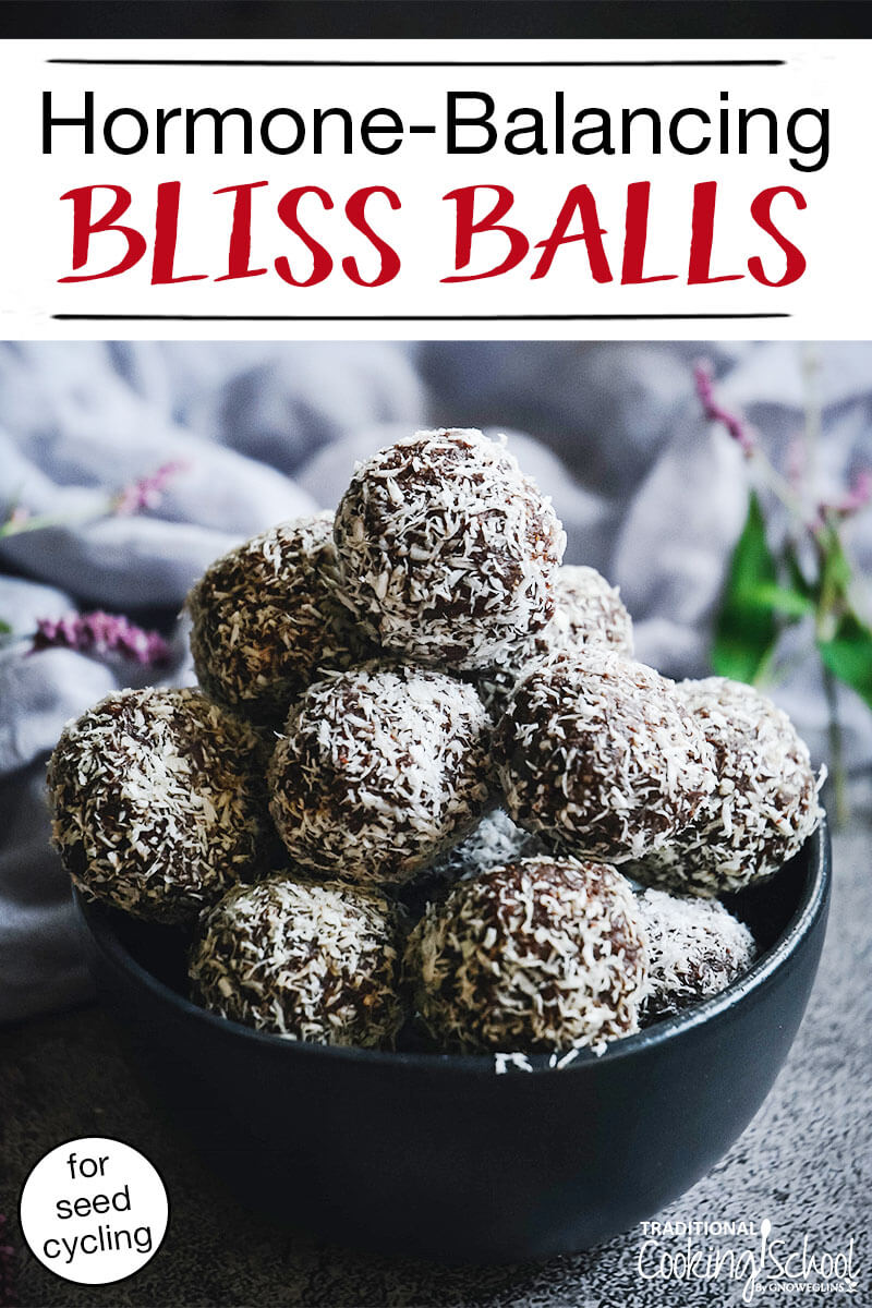 black bowl filled to overflowing with small cacao date balls coated with shredded coconut, with text overlay: "Hormone-Balancing Bliss Balls For Seed Cycling"