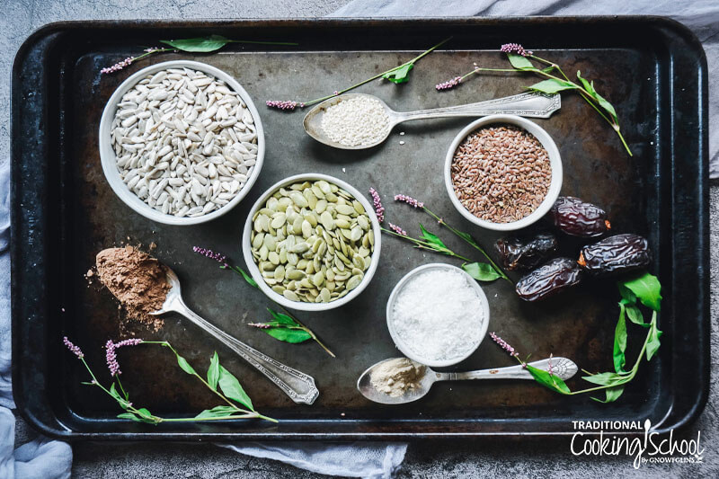 array of ingredients to make cacao date bliss balls spread out on a rustic baking sheet, including spoonfuls of cacao powder, sesame seeds, and maca, as well as dates and bowls of sunflower seeds, pumpkin seeds, flax seeds, and shredded coconut
