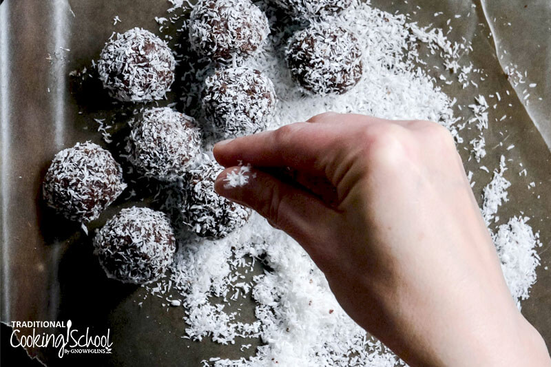 woman's hand sprinkling shredded coconut over an assortment of cacao date bliss balls on a baking sheet