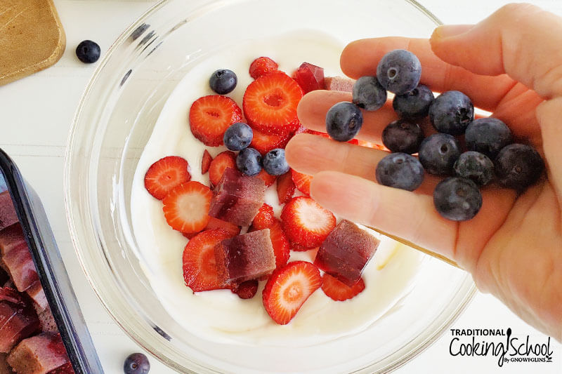 woman's hand pouring a handful of blueberries into a clear glass bowl of berries, homemade jello cubes, and yogurt