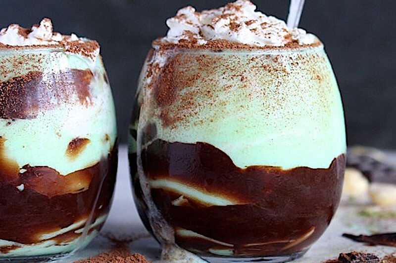 two clear glass cups of an iced vanilla hazelnut chocolate beverage, with whipped cream and cocoa powder sprinkled on top