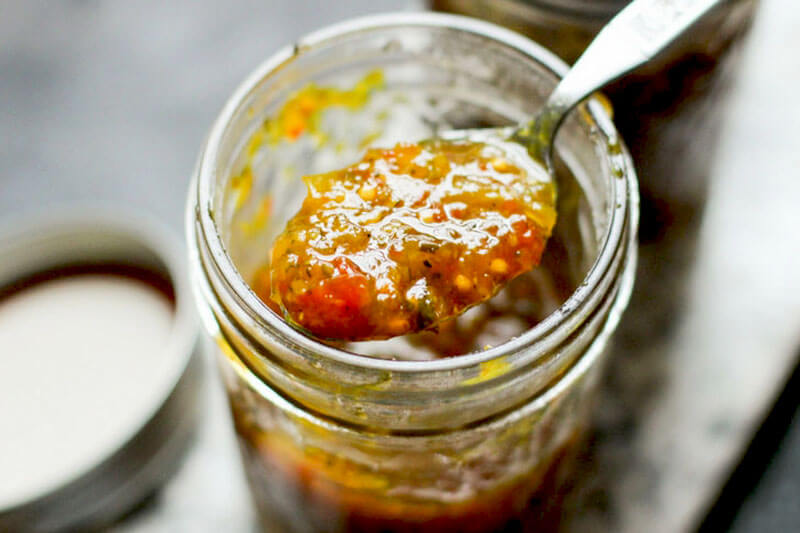small pint-sized Mason jar with a spoon scooping out its contents: orange-yellow colored zucchini relish for hamburgers