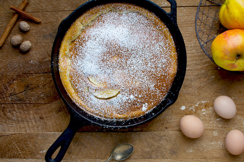 Dutch baby apple pancake in a large cast iron skillet, dusted with sugar on the top, surrounded by ingredients such as cinnamon sticks and whole eggs