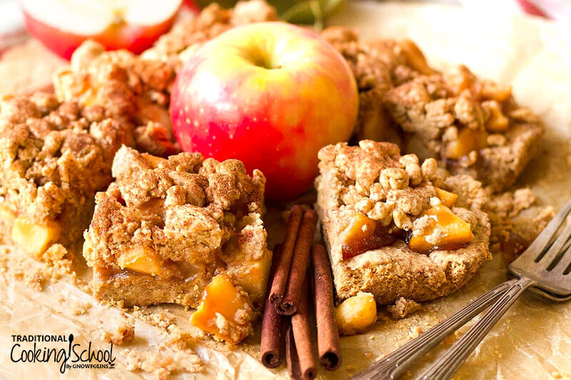 apple pie shortbread bars with a crumble topping, arranged around a whole apple, cinnamon sticks and forks in the foreground