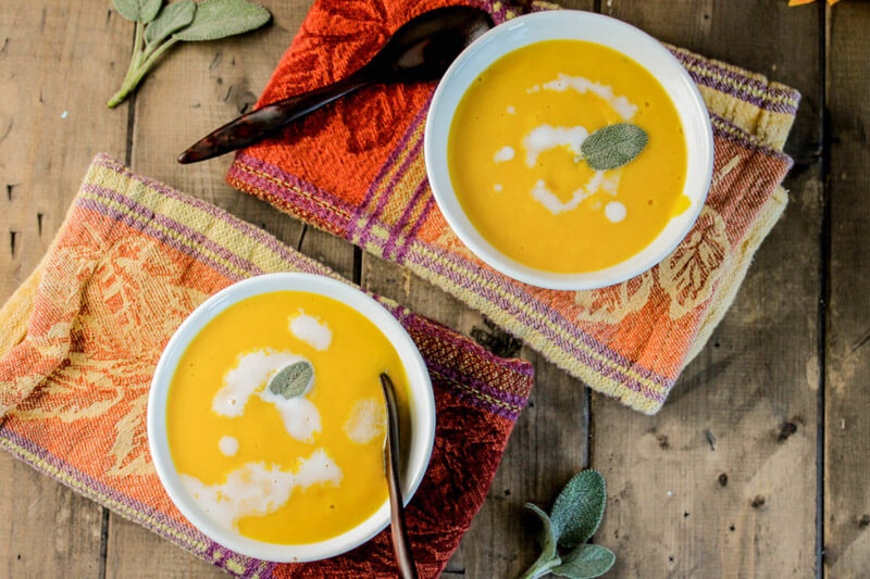 two bowls of creamy pumpkin soup on a wooden table top, garnished with fresh sage and a drizzle of cream