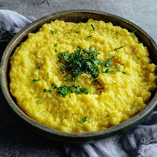 golden-colored creamy polenta in a wooden bowl on a rustic stone backdrop, and fresh green herbs for garnish