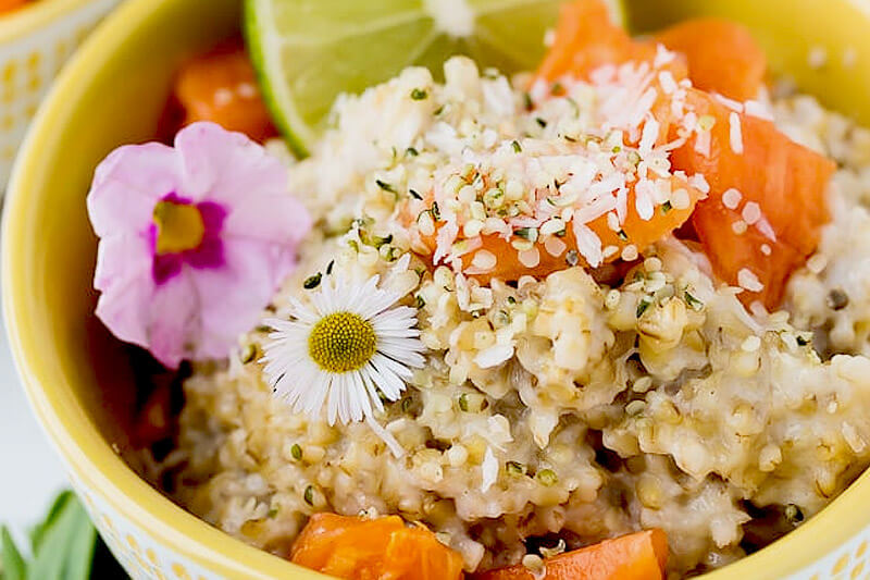 bowl of steel cut oats for breakfast, topped with shredded coconut, mango chunks, a lime slice, and colorful flowers