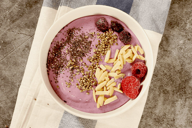 light pink smoothie bowl, topped with chia seeds, hemp hearts, slivered almonds, and frozen berries
