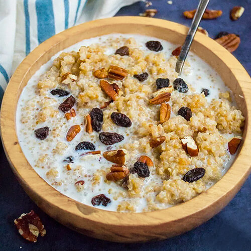 quinoa breakfast porridge, in a wooden bowl with milk and a spoon, topped with raisins and chopped pecans