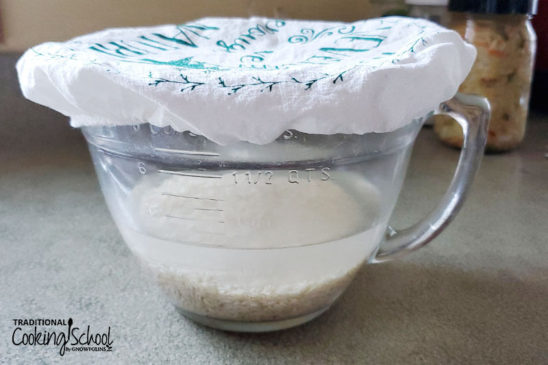 large transparent glass batter bowl of rice that is soaking in water, with a white embroidered cloth over top