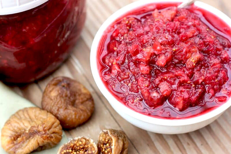 white bowl of fermented cranberry sauce on a wooden background next to a small glass jar of cranberry sauce, and several figs