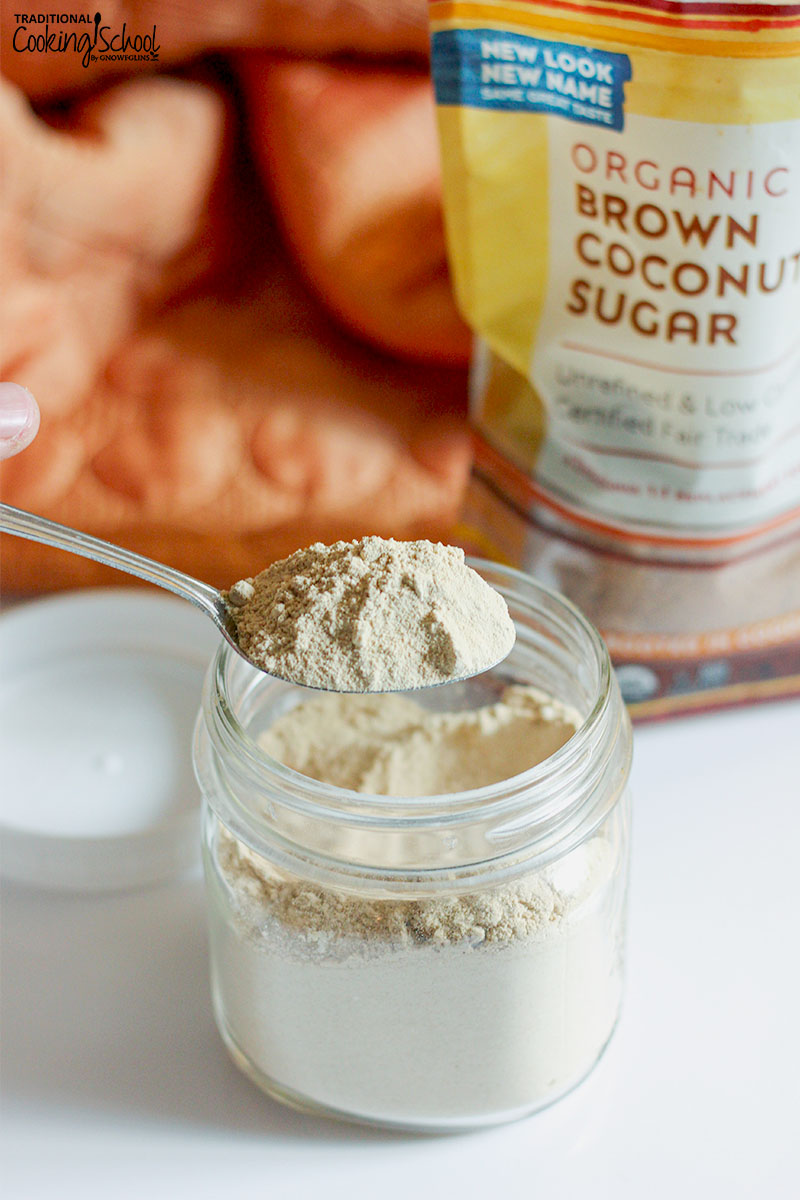 woman's hand holding up a heaping spoonful of finely powdered sugar out of a small jar full of it, with a bag of organic brown coconut sugar in the background