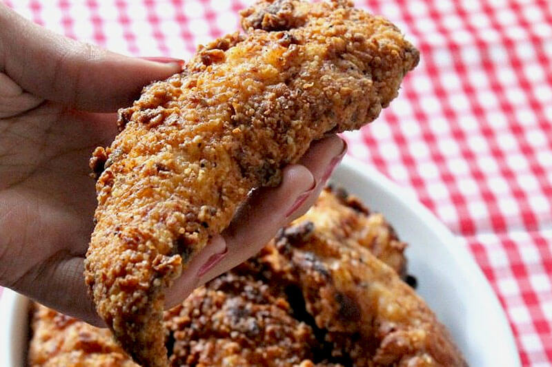woman's hand holding up crispy fried chicken over a white and red checkerboard table cloth, with a ceramic dish of more fried chicken in the background
