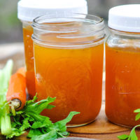 three pint-sized glass jars full of vegetable broth, next to a carrot and fresh herbs