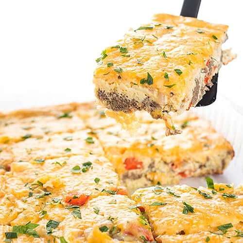 photo of a spatula holding up a slice of cheesy breakfast casserole, with the rest of the casserole in the background