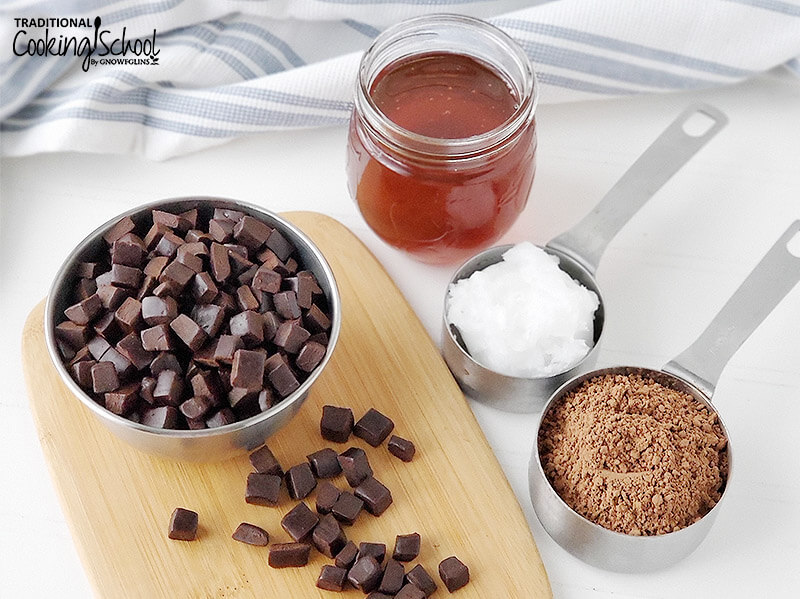 small metal bowl of chocolate chunks on a wooden cutting board next to an array of ingredients necessary to make homemade chocolate, including honey, coconut oil, and cocoa powder