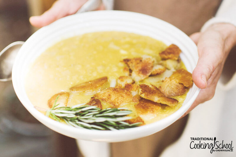 woman's hand holding a large white ceramic bowl of golden-colored, creamy soup topped with chicken and fresh herbs