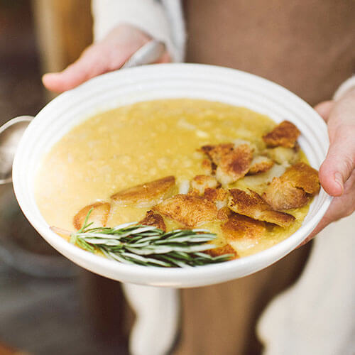 woman's hand holding a large white ceramic bowl of golden-colored, creamy soup topped with chicken and fresh herbs