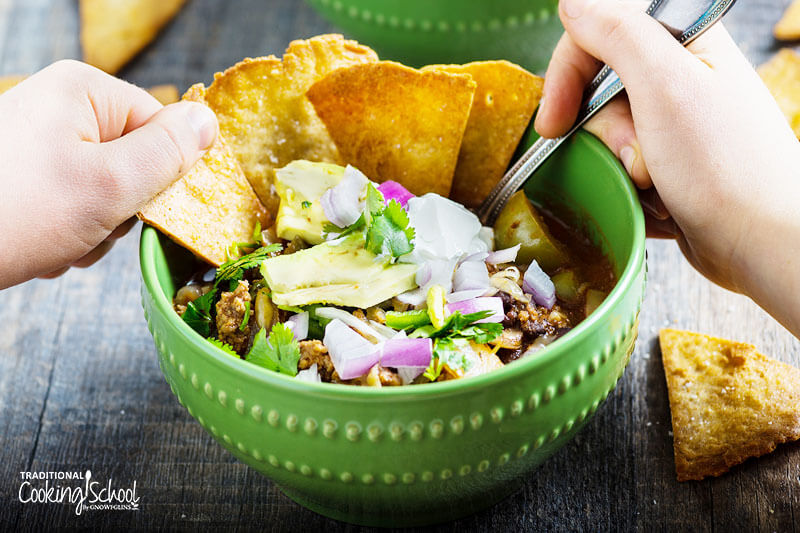 Image of a green bowl filled with chili, topped with red onion, avocado and sour cream. Homemade tortilla chips are dunked in the bowl.